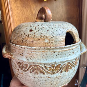 May include: A round ceramic lidded pot with a speckled cream and brown glaze. The pot features two side handles, a curved lid with a central handle, and a cutout for a spoon. A decorative embossed floral vine pattern encircles the main body.