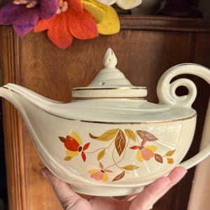 May include: A white teapot with a gold trim and a floral design. The teapot has a curved spout and a handle that curves back towards the spout. The teapot is decorated with a pattern of leaves and flowers in shades of orange, yellow, and red.
