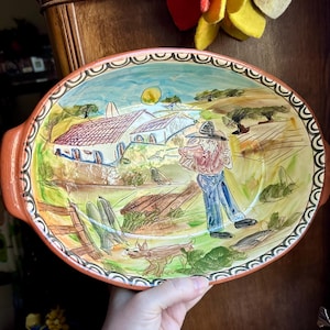May include: An oval ceramic serving dish with a hand-painted scene of a person in a hat and blue jeans walking near a white building with a red roof. The dish features a decorative black and white scalloped border and two side handles.