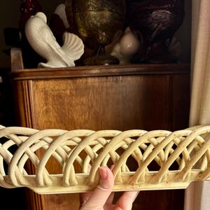 May include: A cream-colored, woven ceramic bread basket. The basket has a long, oval shape with a lattice design. The basket is held by a hand. Other decorative items are in the background.
