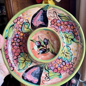 May include: Hand-painted ceramic serving platter with a central dip bowl. The platter features a green rim, with illustrations of grapes, figs, and olive branches in shades of pink, purple, green, and black. The platter is designed for serving appetizers.