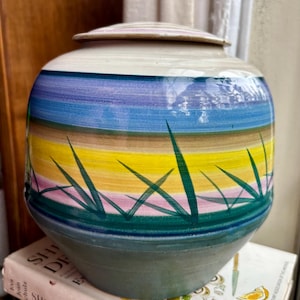 May include: A ceramic jar with a lid, featuring a hand-painted design of horizontal bands in blue, purple, yellow, and brown, with green grass accents. The jar has a rounded shape and a neutral-colored base.