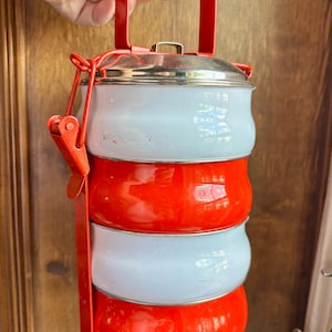 May include: A four-tiered stackable food container featuring alternating light blue and bright orange rounded compartments. The unit is held together by a red metal frame with a top handle and a secure locking mechanism on the side, topped with a lid.
