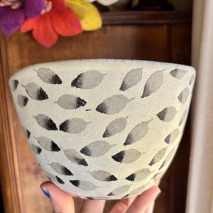 May include: A white ceramic bowl with a black and gray leaf pattern. The bowl is being held by a hand.