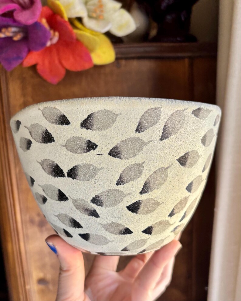 May include: A white ceramic bowl with a black and gray leaf pattern. The bowl is being held by a hand.