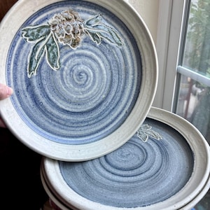May include: A stack of round ceramic plates with a cream-colored rim and a blue spiral pattern in the center. The top plate features a raised, hand-painted floral design in shades of green, brown, and white, showcasing a textured, artisanal finish.