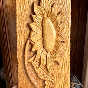 May include: A carved wooden wall art piece featuring a sunflower. The sunflower is the focal point, with detailed petals and a central seed head. The wood has a textured, natural grain, and the piece is rectangular.
