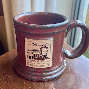 May include: A red ceramic mug with a brown handle. The mug has a rectangular sticker on the front with a black and white image of Abraham Lincoln and the text "Abraham Lincoln Presidential Library & Museum Springfield, Illinois".