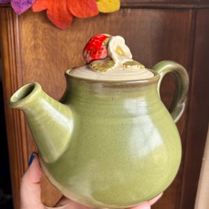 May include: A green ceramic teapot with a strawberry and flower lid. The teapot has a rounded body and a spout on the side.
