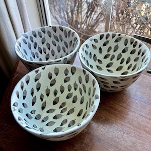 May include: Three white ceramic bowls with a black leaf pattern. The bowls are nested together on a wooden surface.