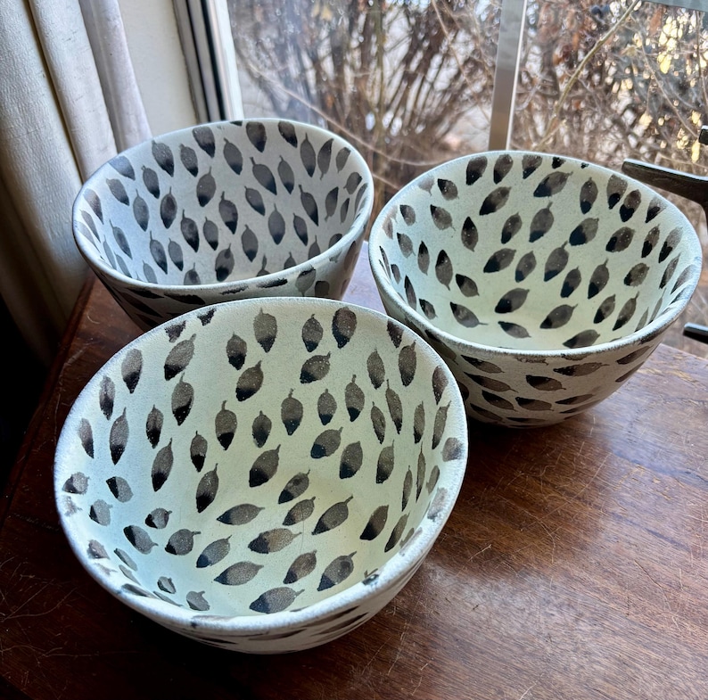 May include: Three white ceramic bowls with a black leaf pattern. The bowls are nested together on a wooden surface.
