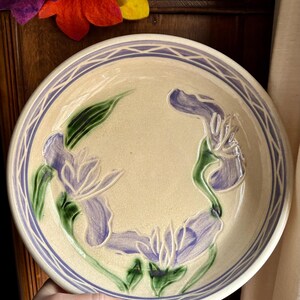 May include: A ceramic serving bowl with a cream-colored glaze and a blue and white geometric border. The bowl features a raised design of purple irises with green leaves.