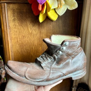 May include: A brown ceramic boot-shaped planter with a cream-colored interior. The boot has silver-colored button details and a lace-up design. The planter is a decorative item.