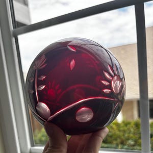 May include: A spherical, deep red glass paperweight featuring intricate, clear-cut floral patterns etched into its surface. The design includes stylized petals, leaves, and circular accents that contrast against the dark, translucent ruby-colored glass.