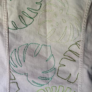 May include: Close-up of a light purple denim jacket back with embroidered green monstera leaves. The leaves are outlined in various shades of green thread, creating a textured, botanical design. The jacket has visible seams and a classic denim construction.