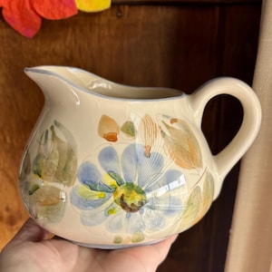 May include: A cream-colored ceramic pitcher with a blue, yellow, and green floral design. The pitcher has a rounded body and a handle.