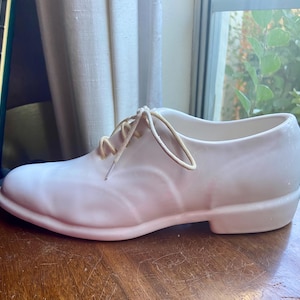 May include: A white ceramic shoe planter with a lace-up design. The laces are a light brown color.