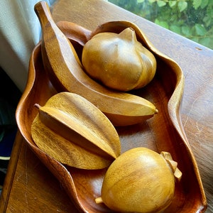May include: A wooden bowl with a carved fruit design. The bowl is filled with four wooden fruit sculptures: a banana, a starfruit, a pear, and a pineapple.