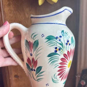 May include: Cream-colored ceramic pitcher with a curved handle and spout. The pitcher is decorated with hand-painted floral designs in red, green, blue, and yellow. A blue stripe accents the rim and base.