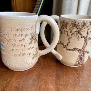 May include: Two handmade ceramic mugs. One mug features a quote by George B. Shaw, with a tree branch design. The other mug has a tree design. Both mugs have a cream-colored base and unique handle shapes.