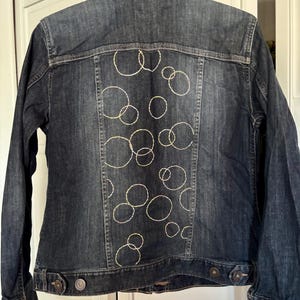 May include: Dark wash denim jacket with gold embroidered circle design on the back. The jacket has a classic collar, button closures, and long sleeves. The denim has a faded, worn look.