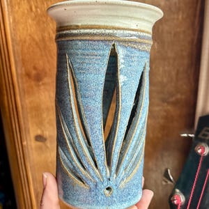 May include: A handmade ceramic vase with a cylindrical shape and a light blue glaze. The vase features cut-out leaf-like designs and a cream-colored rim. The vase is approximately 6 inches tall.