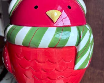 Puede incluir: Un tarro de cerámica para galletas con forma de pájaro rojo que lleva un gorro verde con una pequeña baya roja y una bufanda de rayas verdes y blancas. El pájaro tiene un pico amarillo, ojos negros y patas amarillas, con un patrón festoneado.