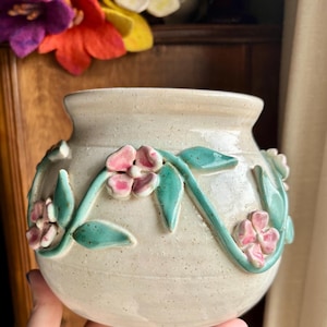 May include: A white ceramic vase with a textured surface and a floral design. The vase is decorated with pink flowers and green leaves.