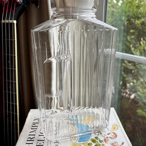 May include: A clear, rectangular glass pitcher with a white lid. The pitcher has vertical ridges and a handle on one side. It is sitting on a book titled "Shrimply Delicious!"
