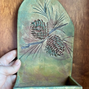 May include: A wall-mounted green and brown ceramic holder with a rounded top. The surface features a relief carving of two pine cones on a branch with needles. The base of the holder has a horizontal row of small, carved pine tree silhouettes.