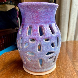 May include: A purple and pink ceramic candle holder with a decorative cut-out design. The holder has a wide base and a narrow opening at the top.
