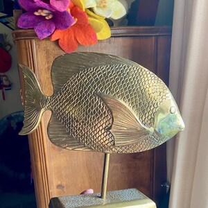 May include: A brass fish sculpture with detailed scale and fin patterns. The fish is mounted on a rectangular base with a slender rod. The sculpture has a golden hue and is a decorative item.