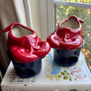 May include: Two ceramic lobster-shaped butter warmers with glossy red tops and dark blue speckled bases. Each piece features a sculpted lobster design with a hollow center and a circular opening at the bottom for a tea light candle.
