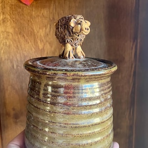 May include: A ceramic jar with a brown and green glaze. The lid is topped with a whimsical ceramic sheep figurine.