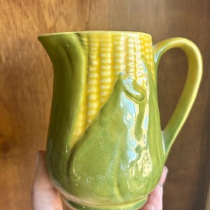 May include: A ceramic pitcher with a corn on the cob design. The pitcher features a yellow corn cob with green husks and a curved handle. The pitcher is likely used for serving beverages and adds a touch of whimsy to the kitchen.