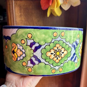 May include: A hand-painted ceramic planter with a vibrant design. The planter features a green and white floral pattern with orange and blue accents. The rim is a deep blue. The planter is oval-shaped and appears to be a decorative item.