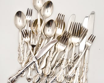 Op de afbeelding: Een verzameling vintage bestek, waaronder lepels, vorken en messen, gerangschikt op een witte ondergrond. Het bestek heeft sierlijke handvatten en een glanzende, reflecterende afwerking, wat wijst op een klassieke stijl.