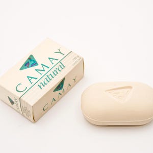 Peut inclure: Un pain de savon blanc naturel Camay et son emballage. Le savon est de forme ovale avec un motif triangulaire. L'emballage est blanc cassé avec le nom de la marque et la description du produit en vert.