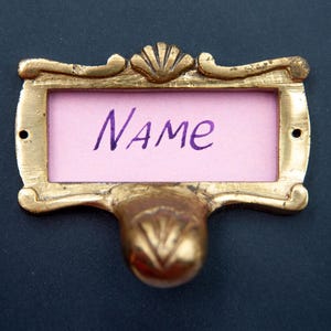 Op de afbeelding: Een gouden metalen naamschild met een roze papieren inzet waarop "NAME" in paarse inkt staat.