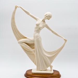 May include: Ivory-coloured Art Deco style sculpture of a woman in a flowing gown, holding draped fabric. The sculpture is mounted on a round, dark wood base. The woman is in a graceful pose, with arms outstretched.