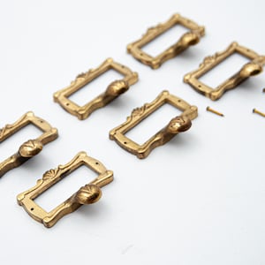 May include: Six gold-toned metal drawer pulls with ornate designs and a small handle. The pulls are arranged in two rows of three. There are also several small gold-toned screws.