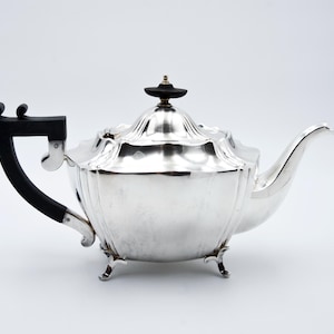 May include: A silver teapot with a black handle and a matching black knob on the lid. The teapot has a curved spout and sits on three small feet. The design is ornate and classic.