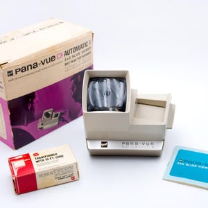 Könnte beinhalten: Ein cremefarbener Pana-vue Automatic 2x2 Diabetrachter mit passender Box und einer kleineren rot-weißen Box. Die Boxen und der Betrachter tragen das Pana-vue-Logo. Eine blaue Karte mit dem Text "2x2 slide viewers" ist ebenfalls vorhanden.