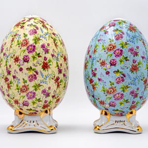 Peut inclure: Deux œufs en porcelaine décoratifs avec des motifs floraux. L'œuf de gauche a un fond jaune avec des fleurs roses et bleues. L'œuf de droite a un fond bleu avec des fleurs roses et blanches. Les deux œufs ont des accents dorés sur la base.