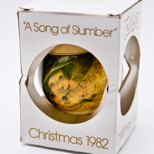 Può includere: Un ornamento natalizio vintage nella sua scatola originale. L'ornamento presenta un bambino che suona la chitarra, con il testo "A Song of Slumber" e "Christmas 1982" stampato sulla scatola. La scatola è bianca con scritte dorate.