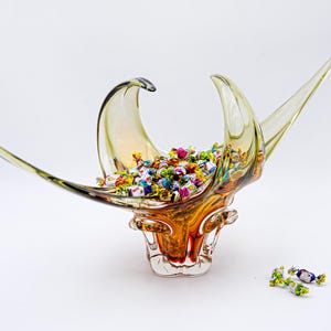 May include: A decorative glass bowl with a unique, abstract design. The bowl is filled with colorful wrapped candies. The glass has amber and clear tones, with long, curved extensions. A few candies are scattered near the base.