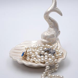 Puede incluir: Un joyero de cerámica blanca con forma de pez con un collar de perlas y un anillo con una joya azul en su interior.