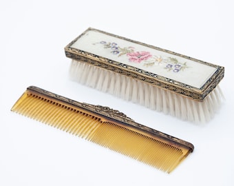 May include: A vintage hair care set with a white brush with a floral embroidered design and a brown comb with gold accents.