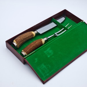 May include: A vintage carving set featuring a knife and fork with antler handles and silver-toned metal accents. The set is presented in a brown box with a green velvet lining, ideal for serving meat.