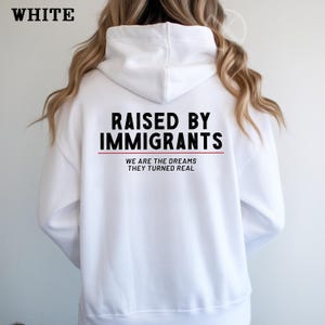 May include: White hoodie with the text "RAISED BY IMMIGRANTS" in black, with a red line underneath. Below the line, the text reads "WE ARE THE DREAMS THEY TURNED REAL". The word "WHITE" is in the top left corner.